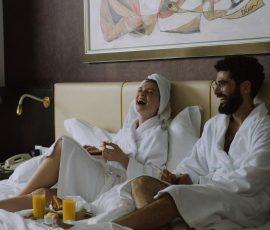 Europe A joyful couple in bathrobes laughing while having breakfast in bed indoors.
